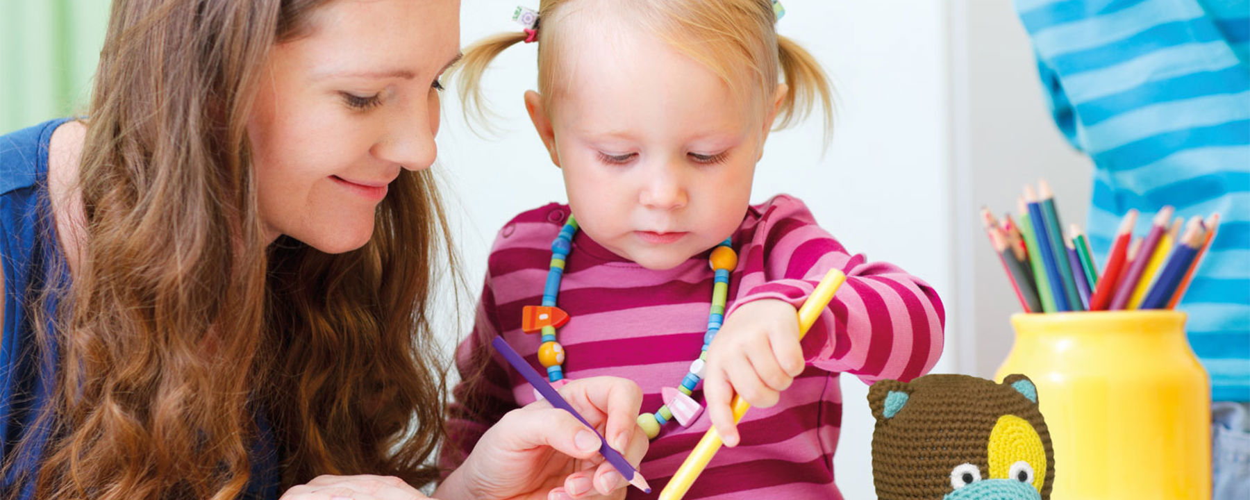 En mamma och ett litet barn ritar med kritor tillsammans. Nallen Tryggve sitter i förgrunden.