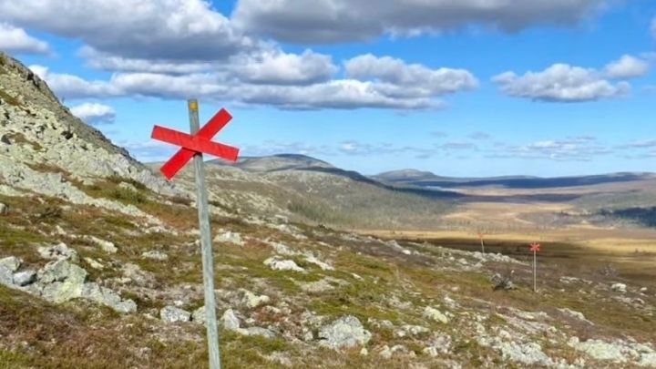 Från svenska fjällvärlden med fjäll i fjärran. Stolpe med rött kryss i förgrunden. Blå himmel med moln.