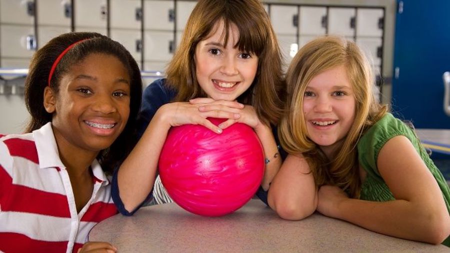 Tre flickor som lutar sig över ett bord. Den mittersta har ett rosa bowlingklot i händerna.