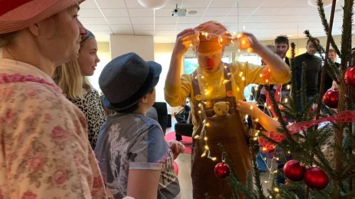 Tre personer titta på en clown som trär en ljusslinga över huvudet. I högerkanten av bilden skymtar en julgran.