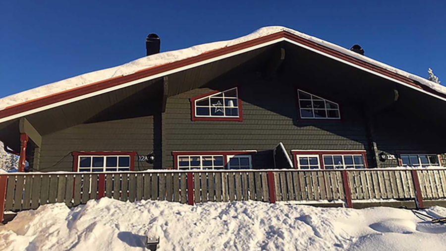 Mörkgrå fjällstuga med snö på taket och framför, i solsken